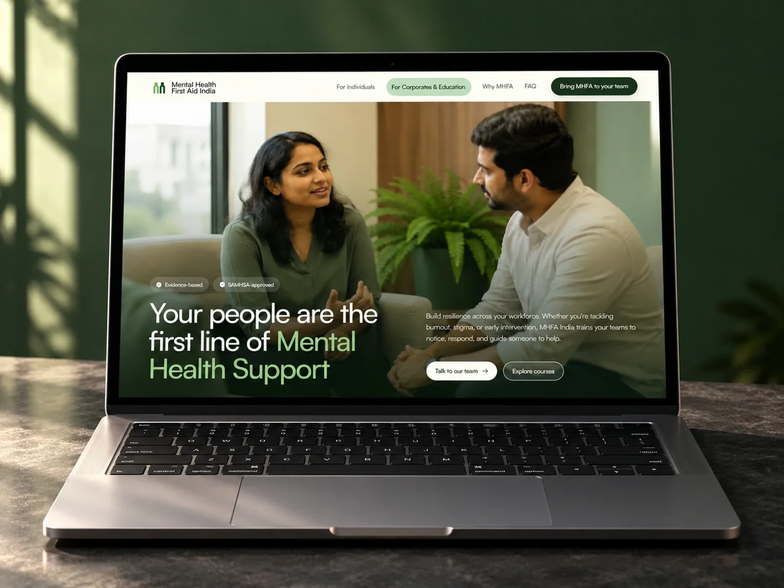 Mental Health First Aid India website on a laptop: "Your people are the first line of Mental Health Support" over two colleagues talking, with Evidence-based and SAMHSA-approved badges